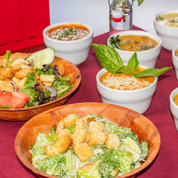 Assortment of Italian soups and salads, including tomato soup with basil and Caesar salad, on a red tablecloth.