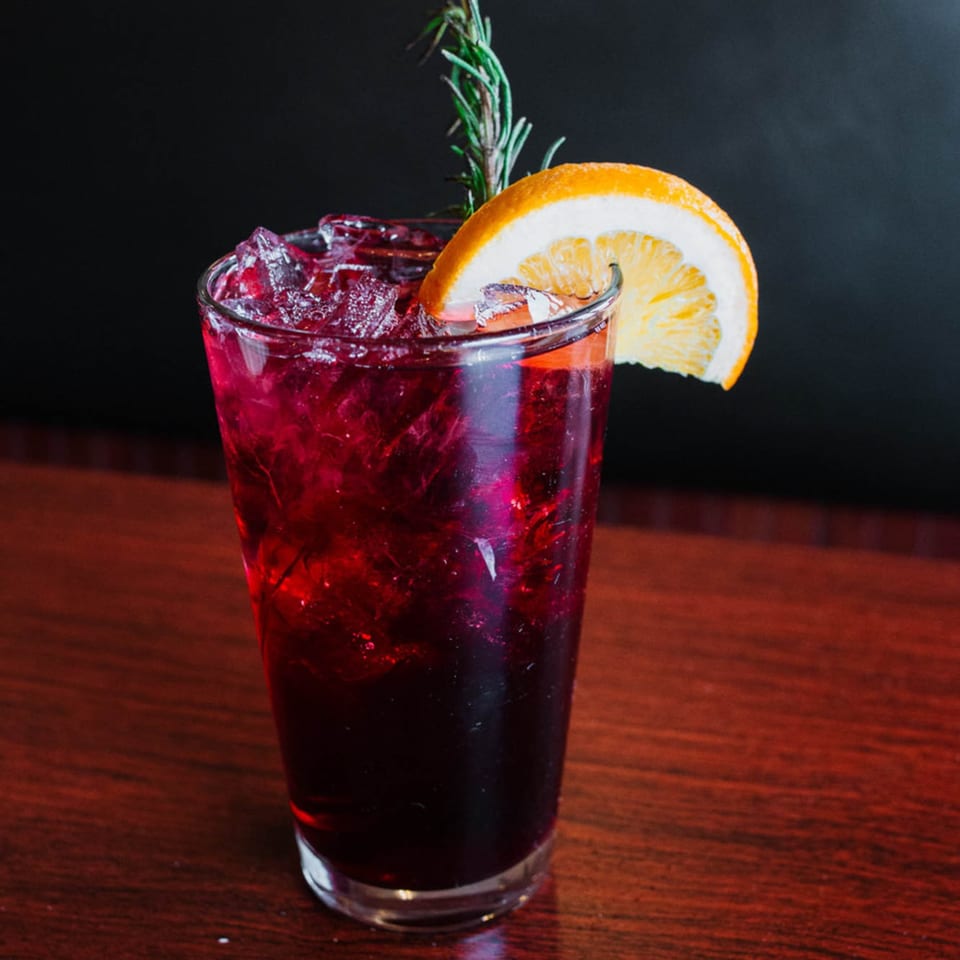 A vibrant red cocktail with ice, garnished with a fresh orange slice and a sprig of rosemary.