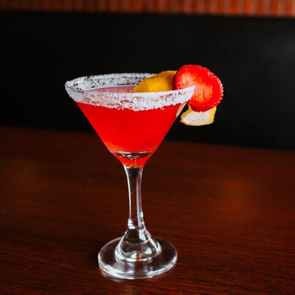 A red martini with a sugared rim, garnished with a strawberry and lemon peel on a dark wooden table.