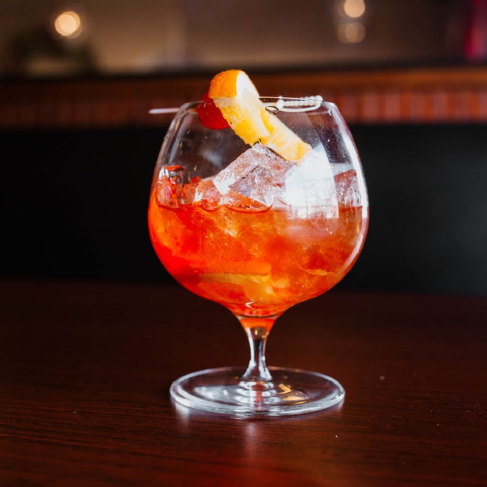 A vibrant orange cocktail with ice, garnished with an orange slice and cherry, on a dark wooden table.