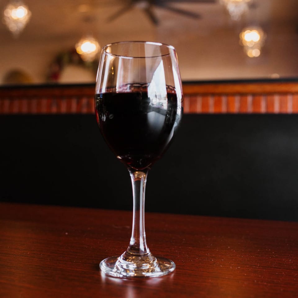A glass of red wine sits on a wooden table at Sorrento Restaurant, with warm lighting in the background.